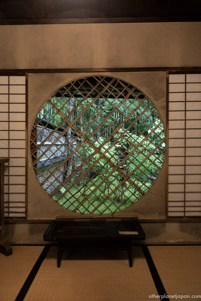  View through the round Yoshino window inside the thatched hermitage at Gioji Temple, framing the moss garden beyond a latticed screen