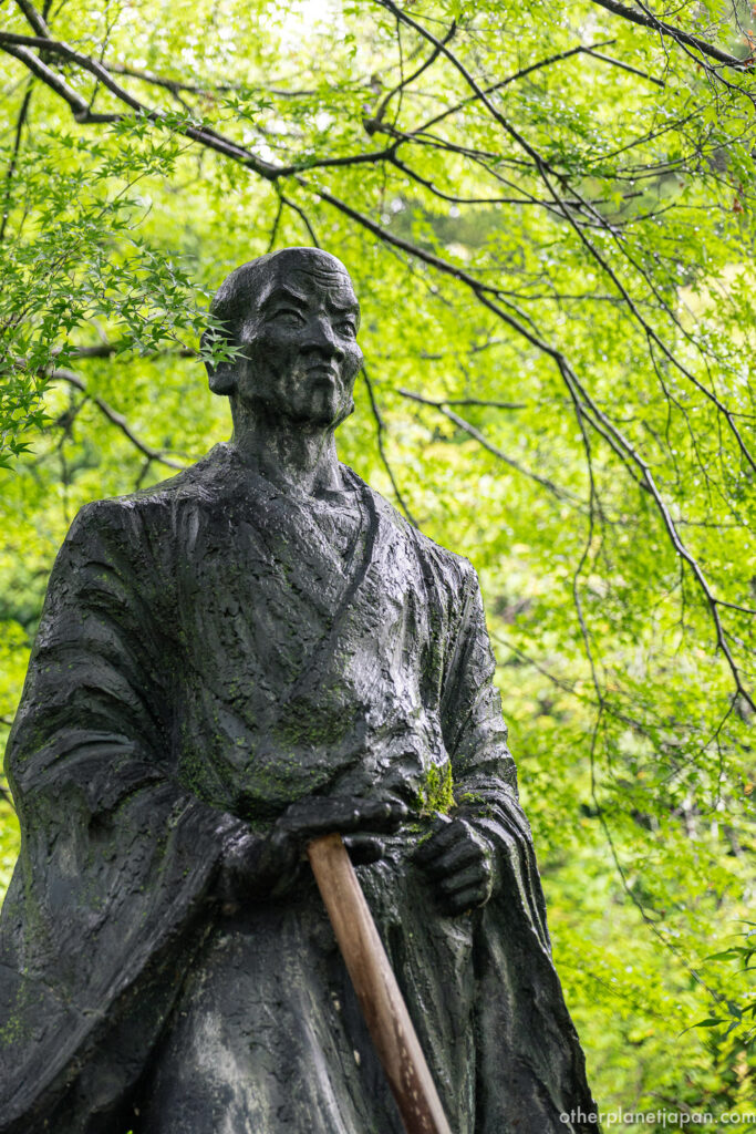 Bronze statue of Suminokura Ryoi holding a staff beneath green maple canopy at Nison-in Temple