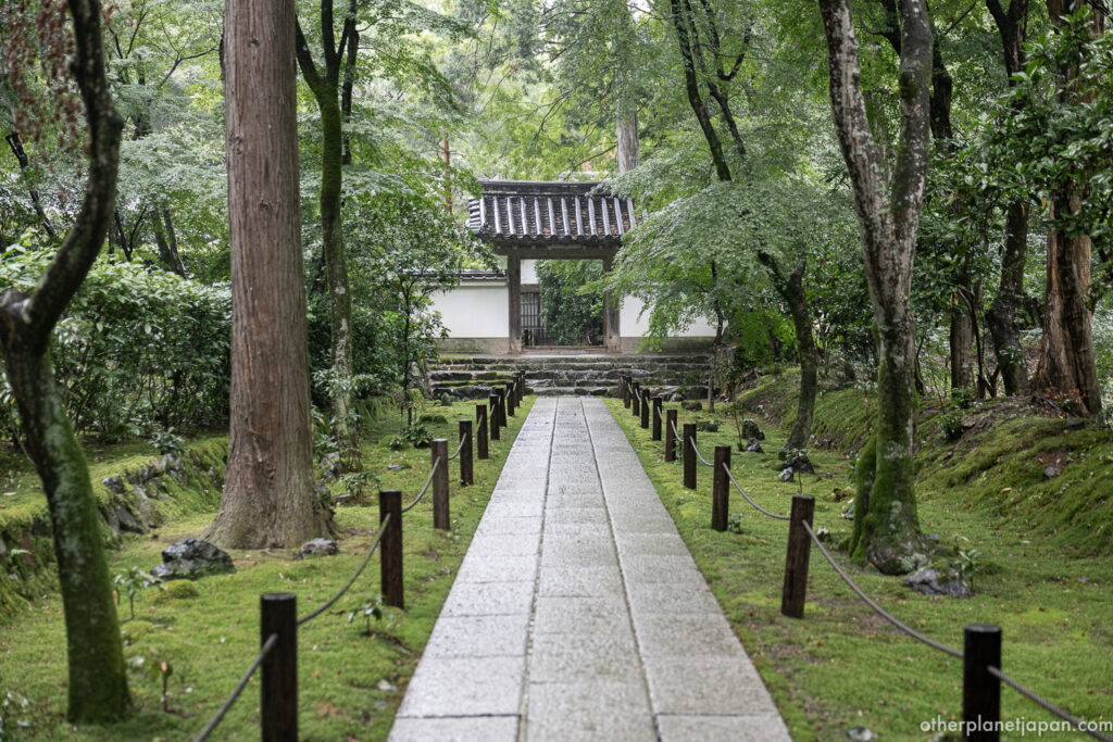 A stone-paved approach flanked by rope-post fencing leads through lush green moss to a white-walled gate at Jizō-in Temple in Kyoto