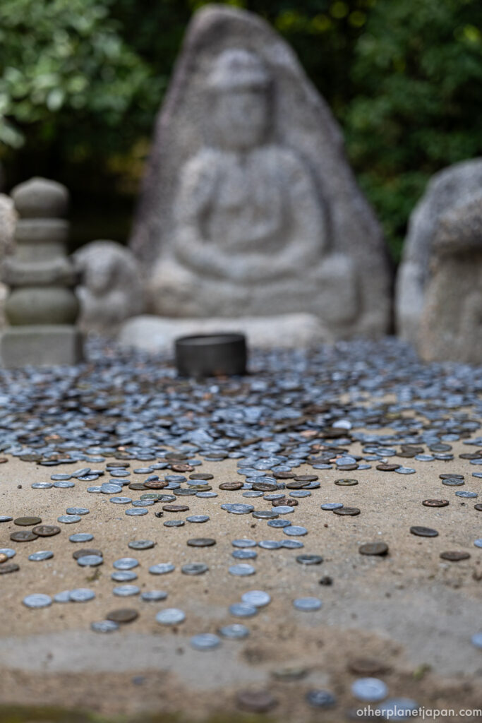 Stone Buddha statue surrounded by scattered coins left by visitors along the garden path at Kinkakuji, Kyoto