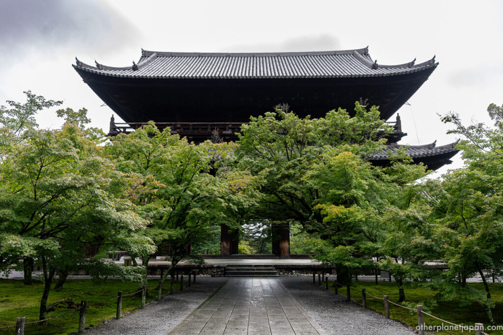 The massive two-tiered Sanmon gate of Nanzenji temple rises above fresh green maple trees in Kyoto, framed by stone paving.