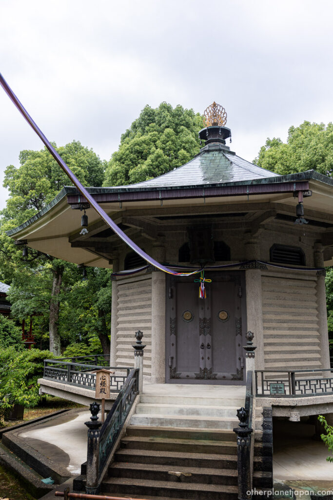 The octagonal Shingyo Hoto pagoda at Daikakuji Temple connected by a purple cord, surrounded by green trees on an overcast day