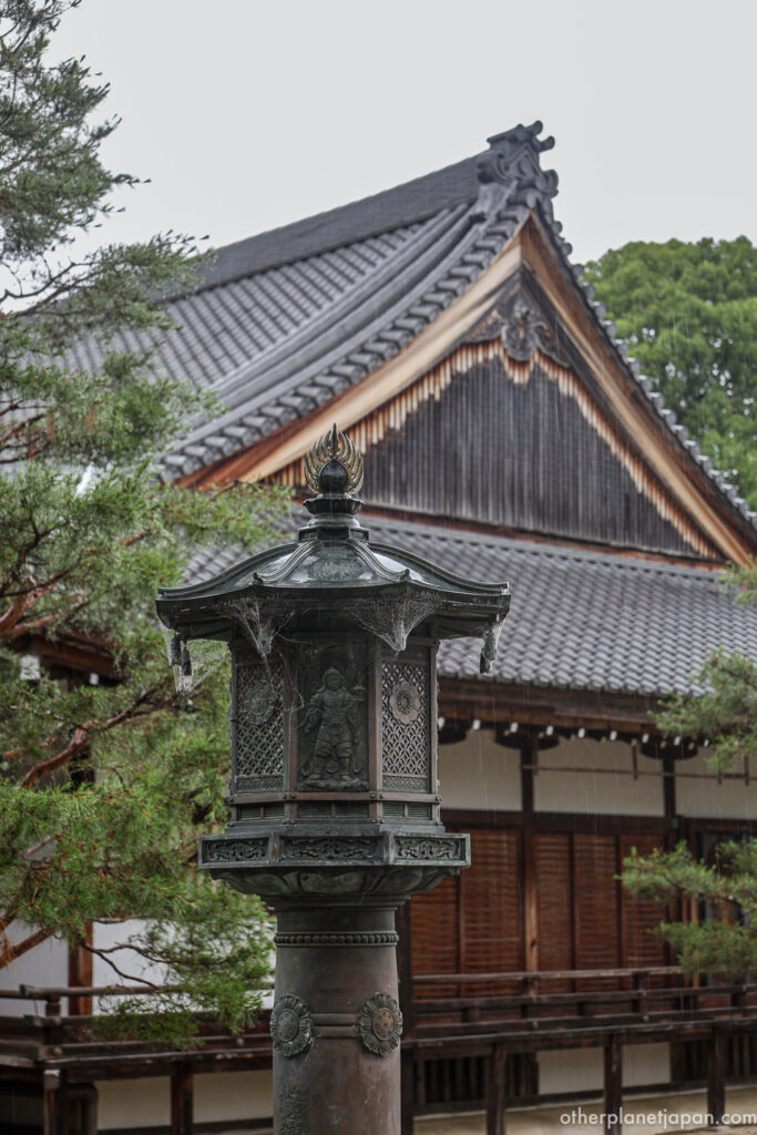 An ornate bronze lantern with carved guardian figures in front of a traditional temple roof at Daikakuji in the rain