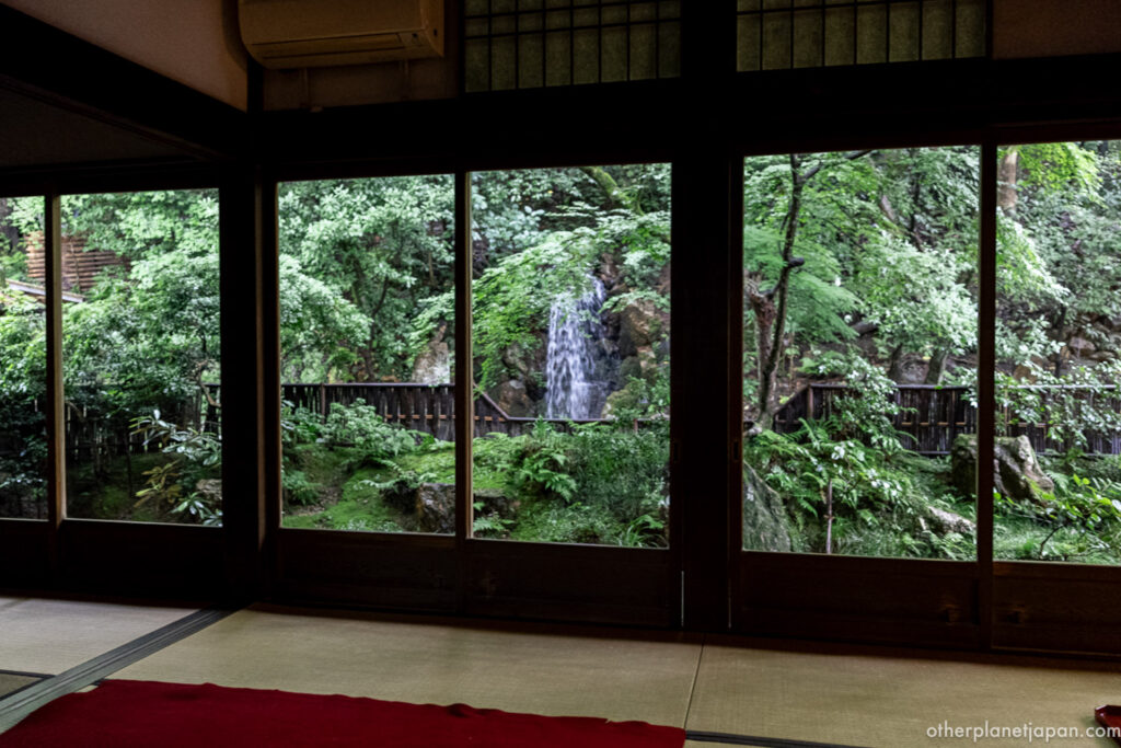 The tatami tea room inside Nanzenji Hojo, with floor-to-ceiling glass doors framing a view of the inner garden's waterfall and dense green foliage in Kyoto.