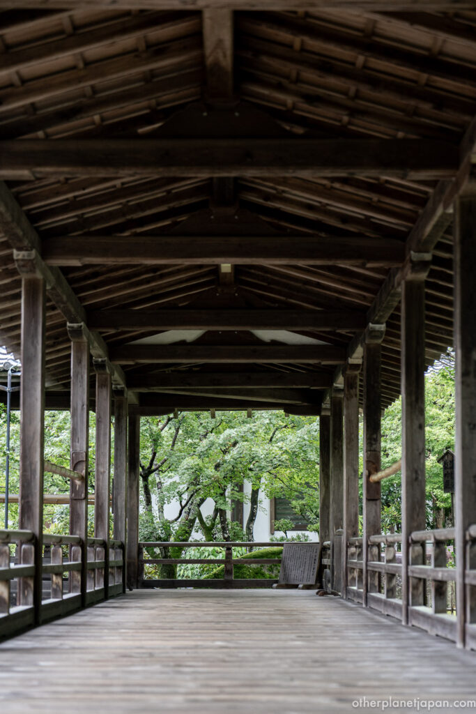 A long covered wooden corridor connecting buildings of Nanzenji's Hojo, leading toward a small framed view of green maples in Kyoto.