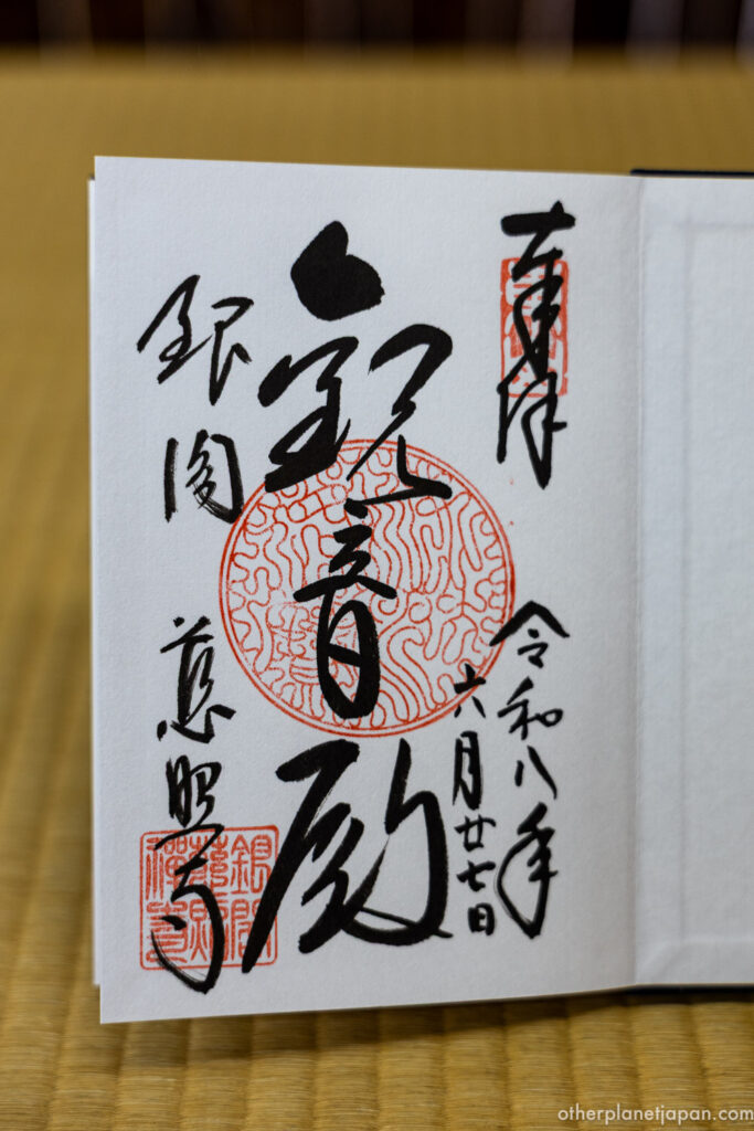 An elegant goshuin (temple stamp) from Ginkakuji, the Silver Pavilion, resting on a tatami mat. The calligraphy reads 'Kannon-den' over a large red seal, capturing the serene atmosphere of a rainy morning at the temple.