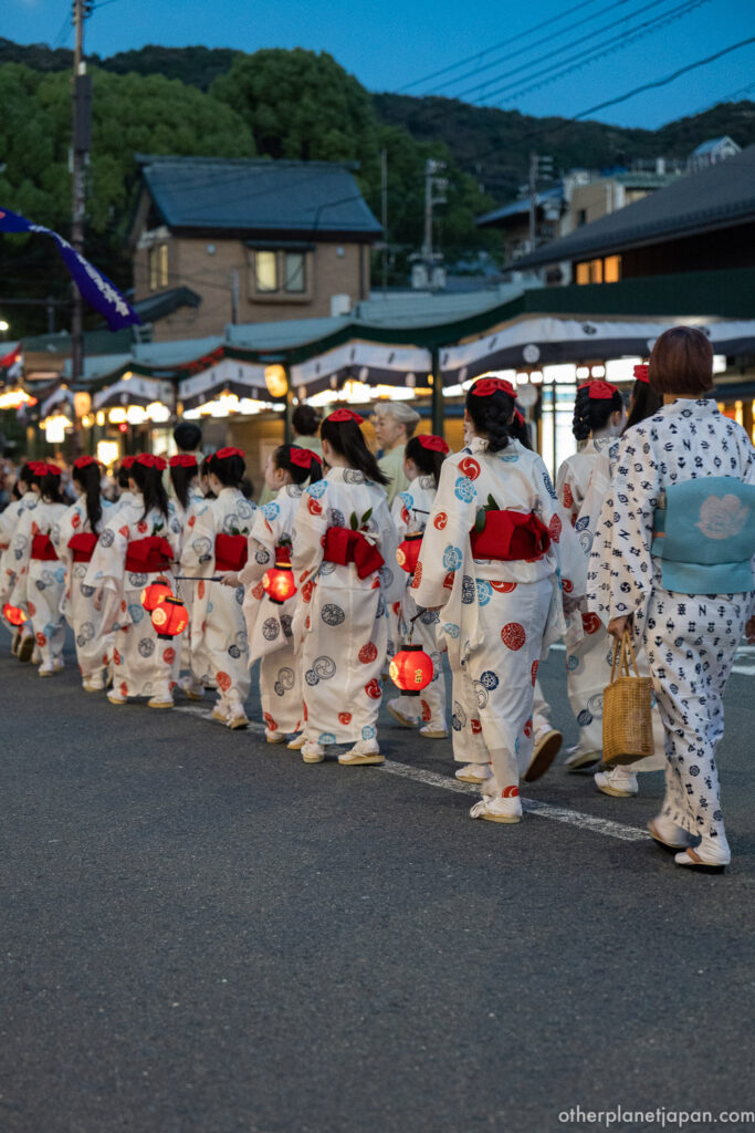 A line of children in white-and-red yukata carrying glowing red lanterns through Kyoto at dusk