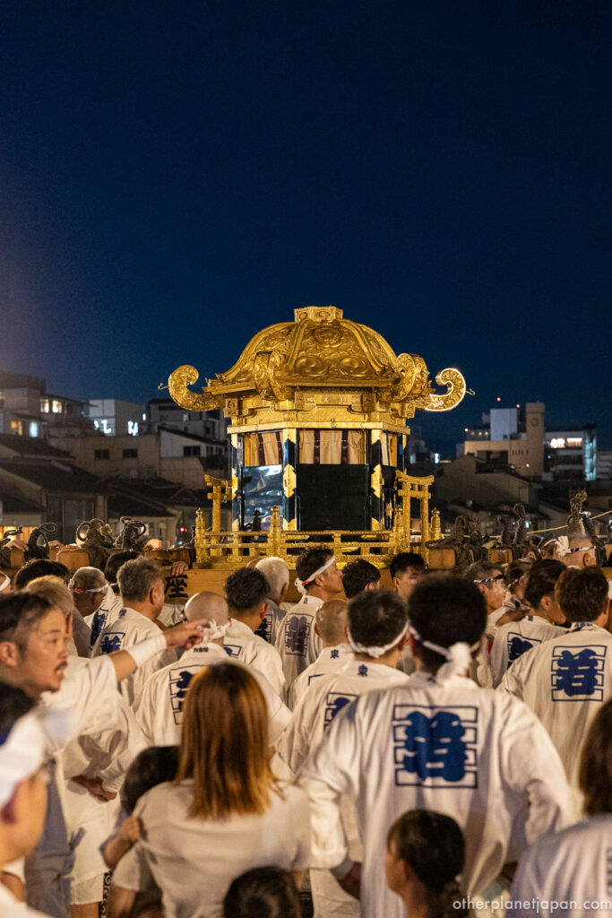 The golden mikoshi surrounded by bearers in white as it moves through the crowd at night