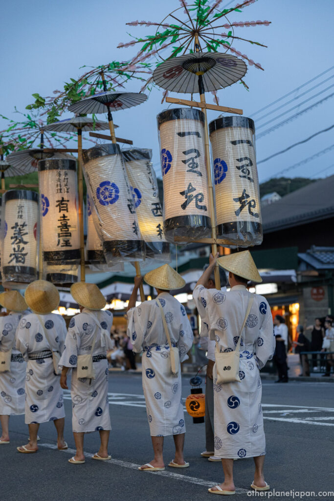 Lantern bearers in white yukata holding tall paper lanterns crowned with decorated umbrellas at dusk