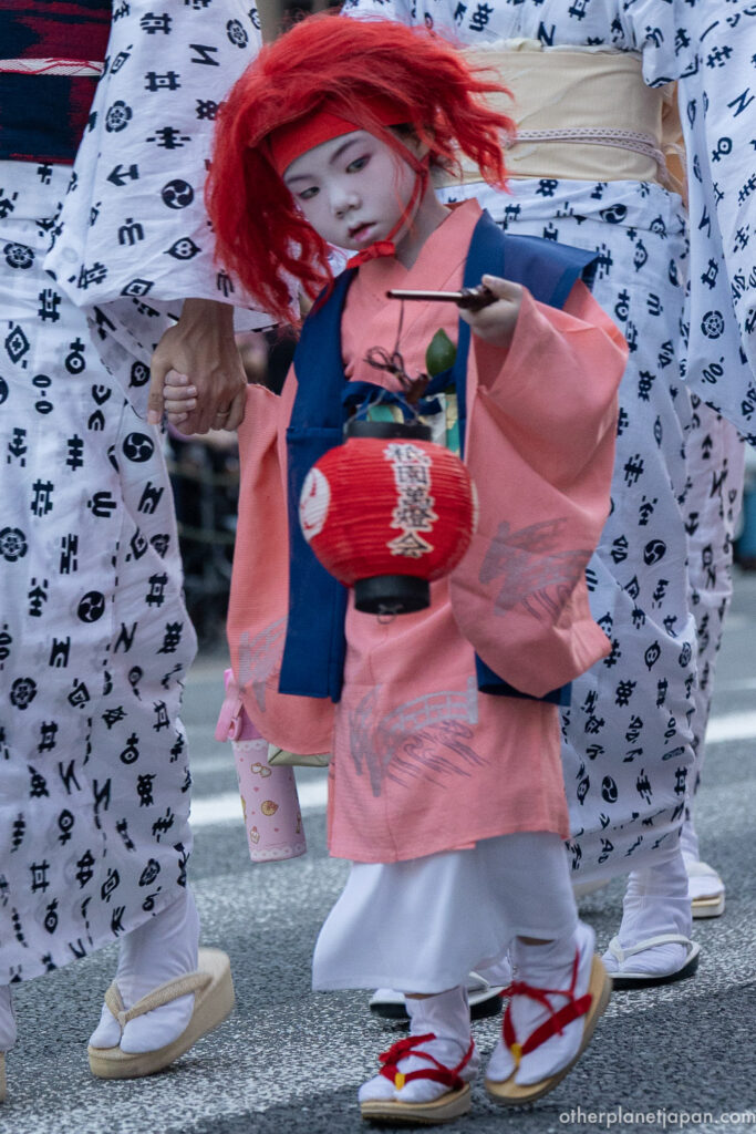 A child wearing the flame-red shaguma wig and white face paint, carrying a lantern in the Omukae Chochin procession