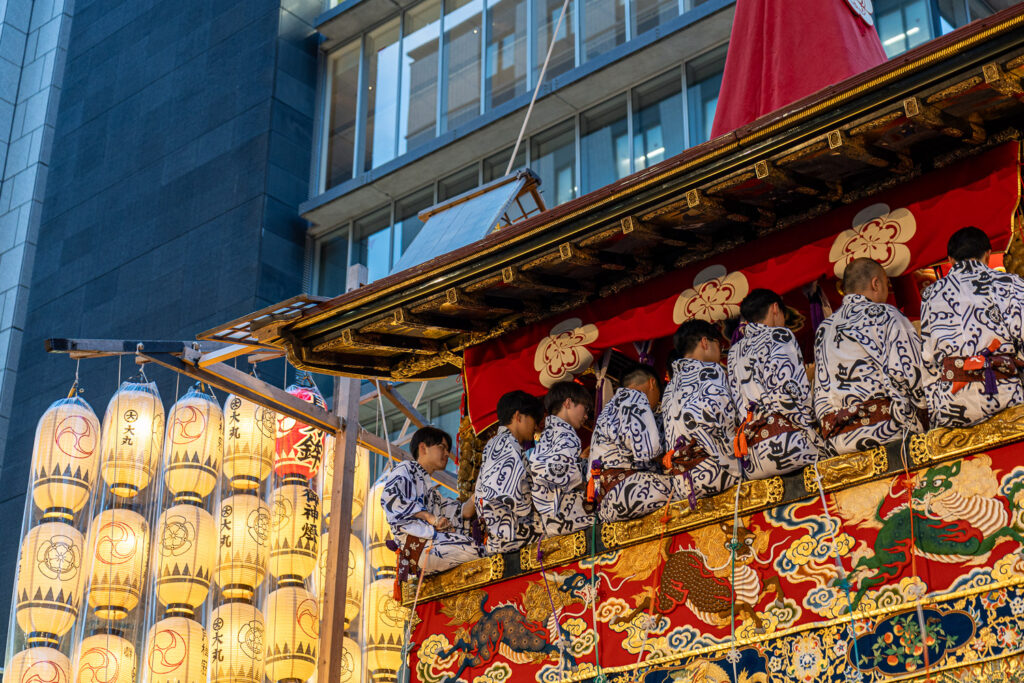 Musicians in yukata seated on an illuminated Gion Matsuri float beneath rows of glowing paper lanterns