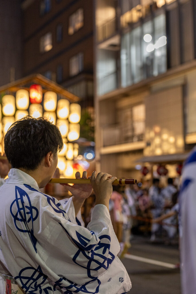 A young flute player in white and indigo yukata seen from behind with festival lanterns glowing in the background