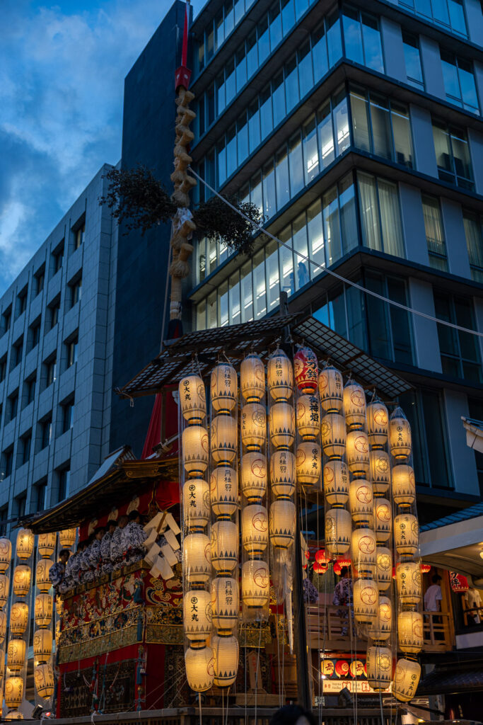 A Gion Matsuri float with sacred rope and lantern rows at twilight against modern Kyoto office buildings