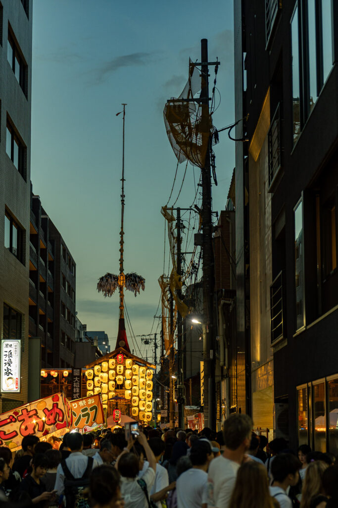 Crowds and food stalls on a narrow Kyoto street at dusk with an illuminated Gion Matsuri float in the distance