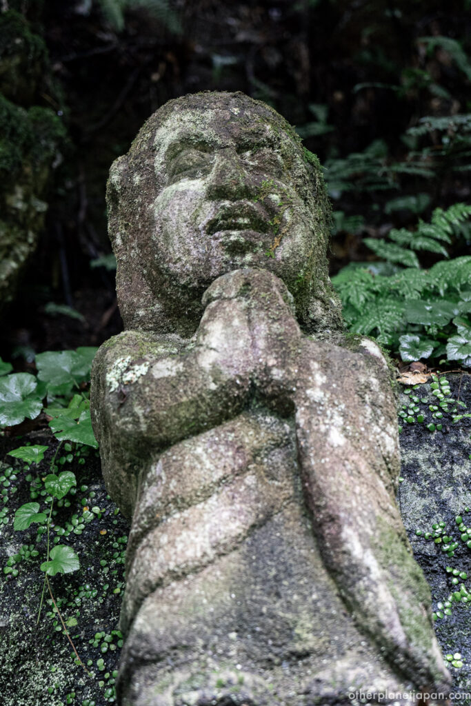 Weathered praying rakan statue with clasped hands surrounded by ferns at Otagi Nenbutsuji, Kyoto