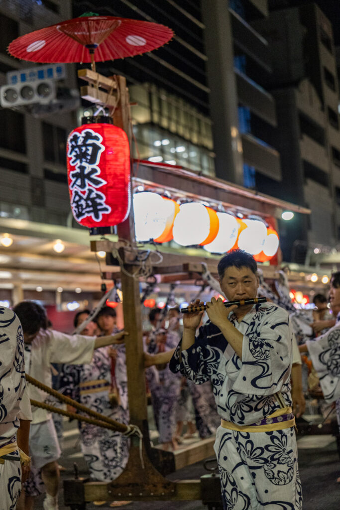 A musician in yukata playing a bamboo flute beneath the red Kikusui Hoko lantern at night during Gion Matsuri