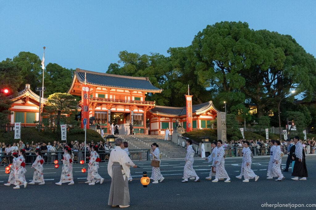 The Omukae Chochin lantern procession passing in front of the illuminated west gate of Yasaka Shrine at dusk, with children and adults in matching yukata carrying glowing paper lanterns during Gion Matsuri on July 10