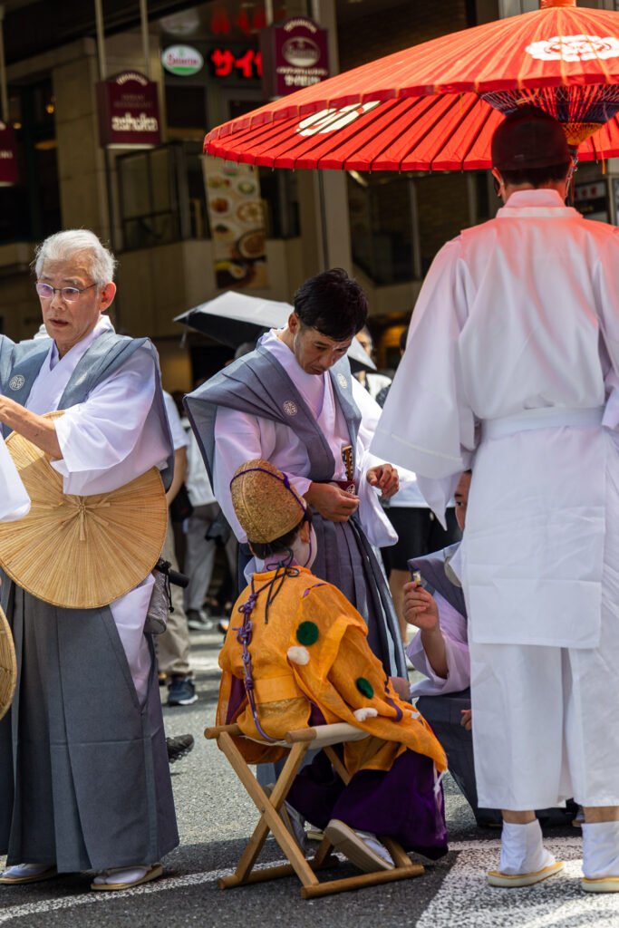 The chigo of Naginata Hoko resting on a folding stool beneath a red parasol