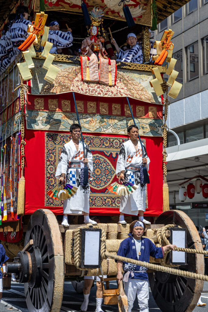 Two men on the front platform of Tsuki Hoko beside its towering wooden wheels