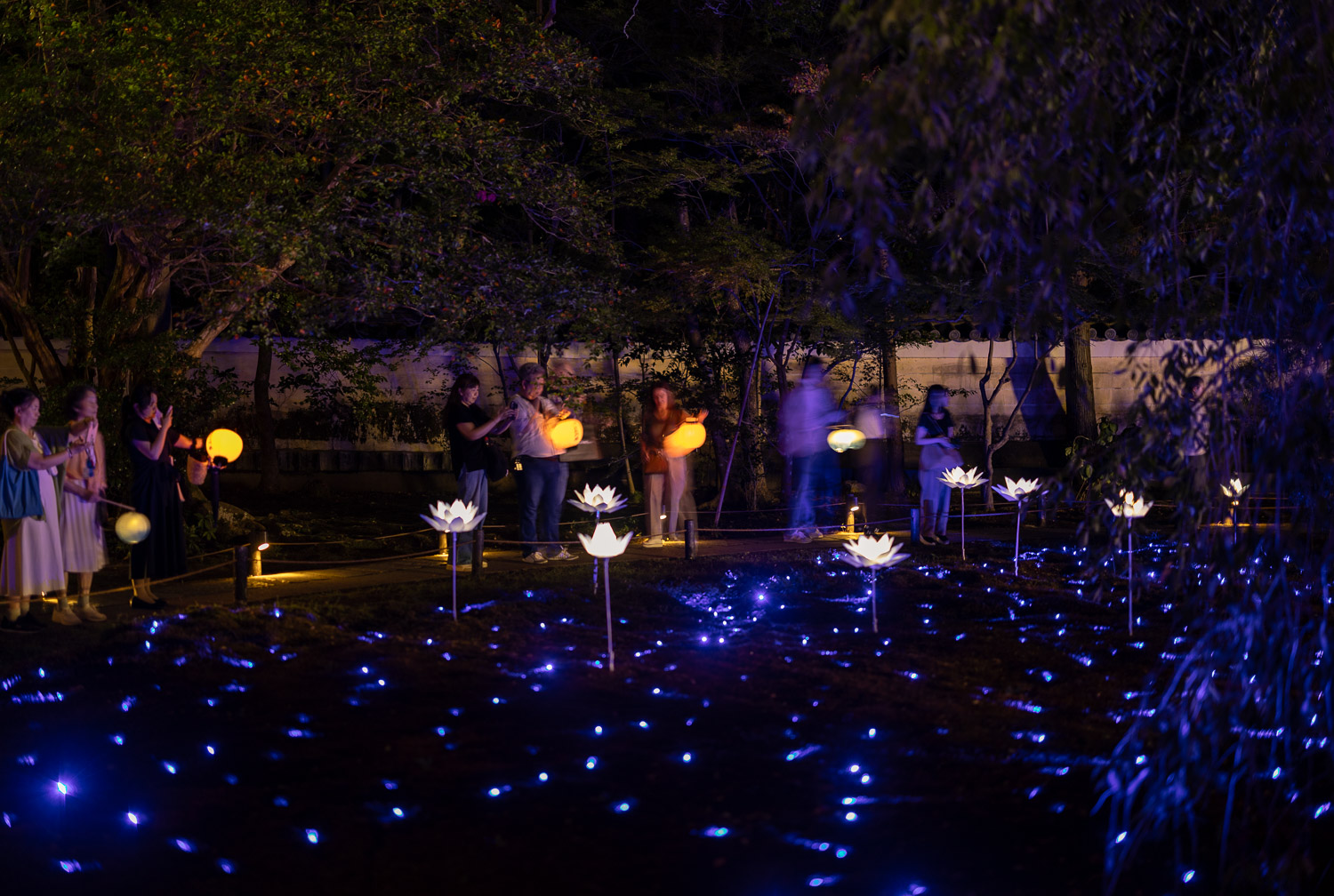 Visitors carrying paper lanterns beside the blue-lit moss garden at the Shoren-in Temple night illumination