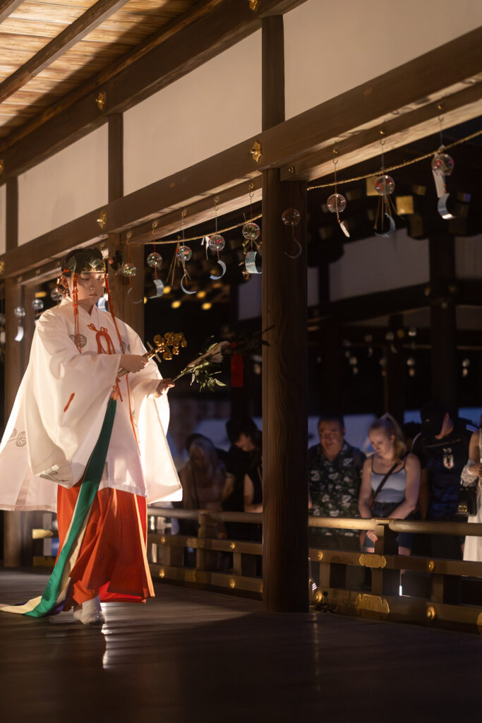 A shrine maiden performing the Kamo-no-Mai kagura dance at the Hashidono hall while international visitors watch from the railing