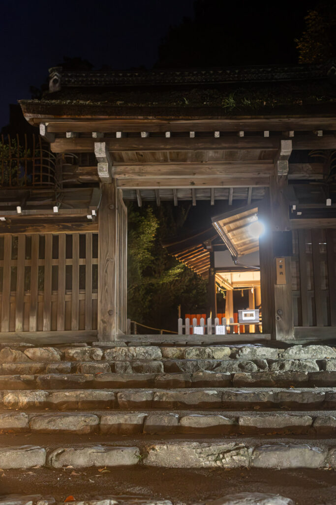 Stone steps leading to the open gate of Shingu Shrine at night, with the inner sanctuary glowing in warm light