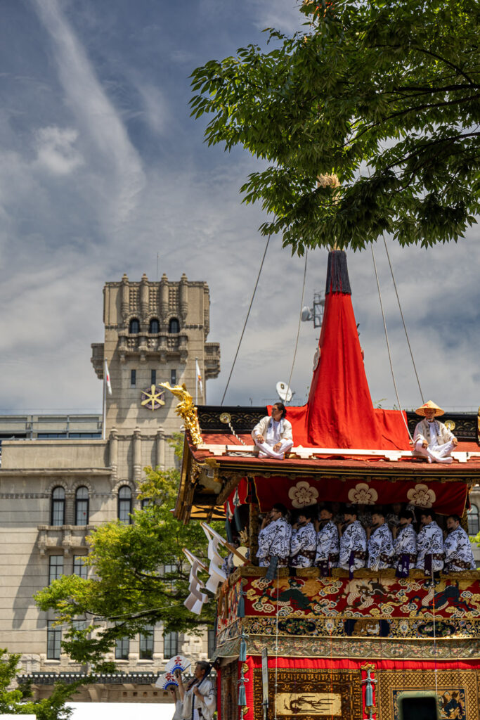 Roof riders on a Gion Matsuri float beside the stone facade of Kyoto City Hall