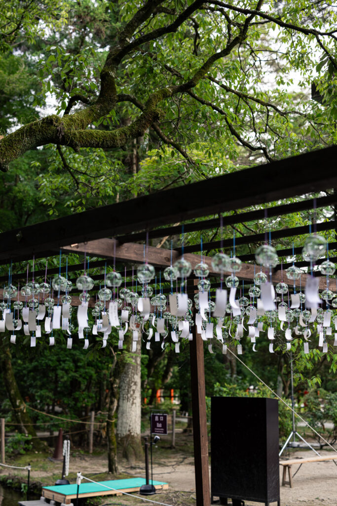 Glass wind chimes with paper strips hanging from a wooden frame under green trees at Kamigamo Shrine