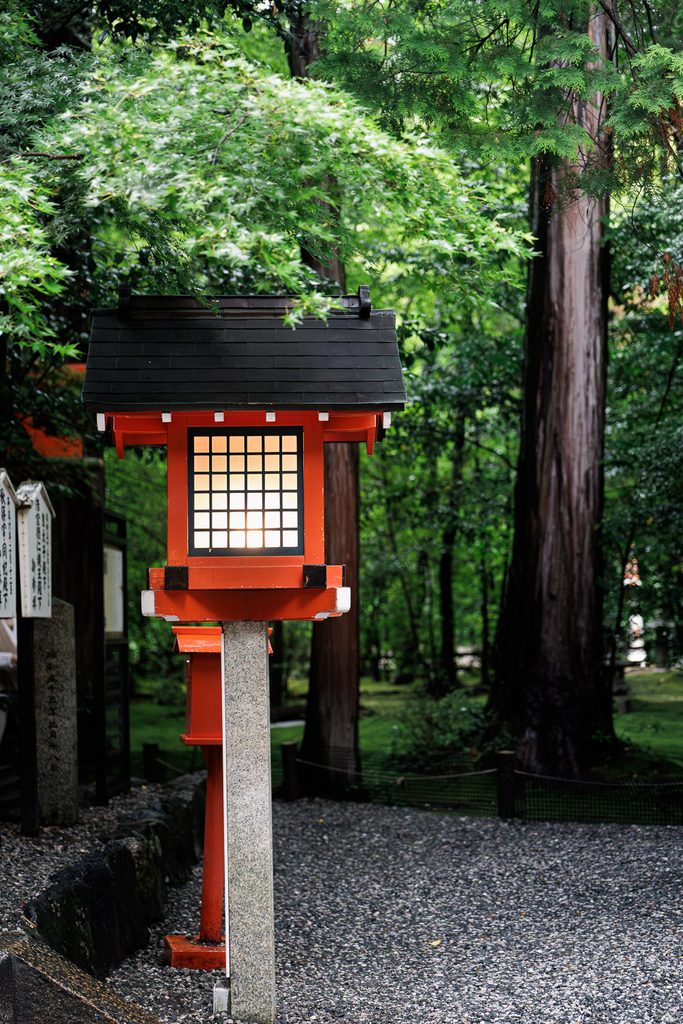 A red wooden lantern with a glowing paper window beside the gravel path at Nonomiya Shrine