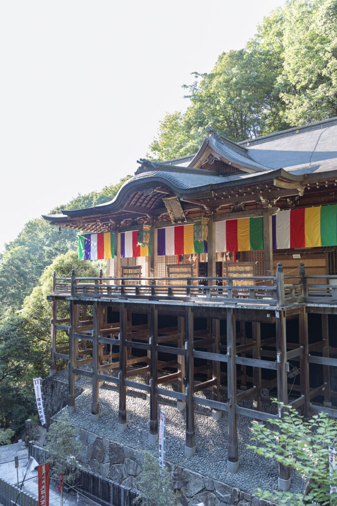 Main hall of Tanukidani Fudoin built on tall wooden stilts against the mountainside, with five-colored Buddhist banners