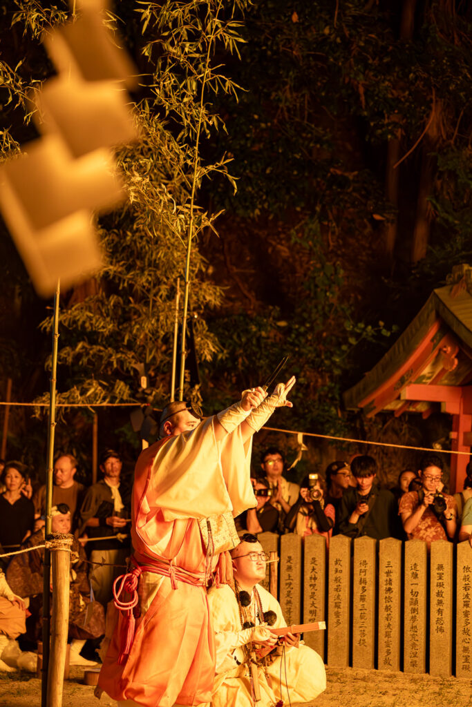 Yamabushi monk pointing skyward during the fire ceremony while spectators watch behind stone pillars at Tanukidani Fudoin