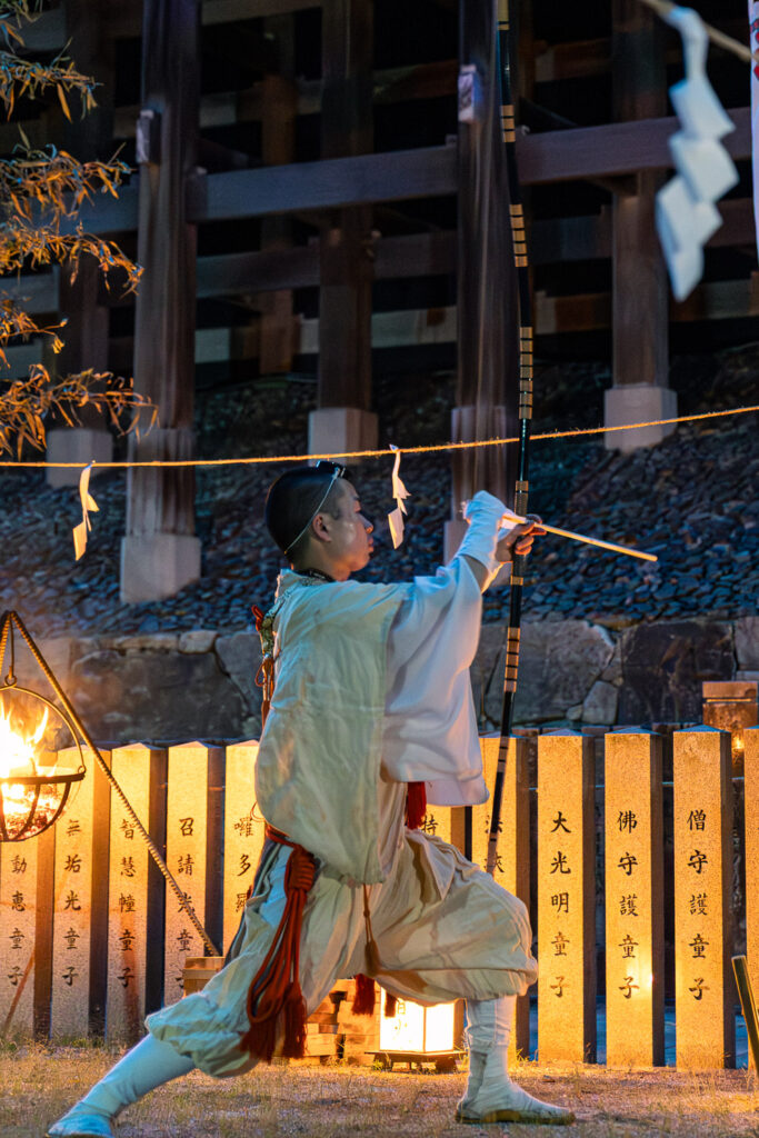 Yamabushi monk in white robes drawing a long bow during the purification ritual at the Tanukidani Fudoin fire walking festival