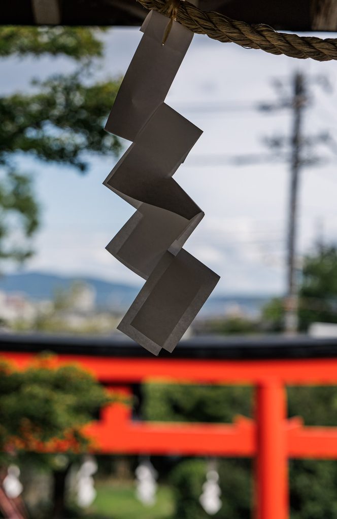 A white paper shide streamer hanging at Tsukiyomi Shrine, with a vermilion torii gate blurred behind it