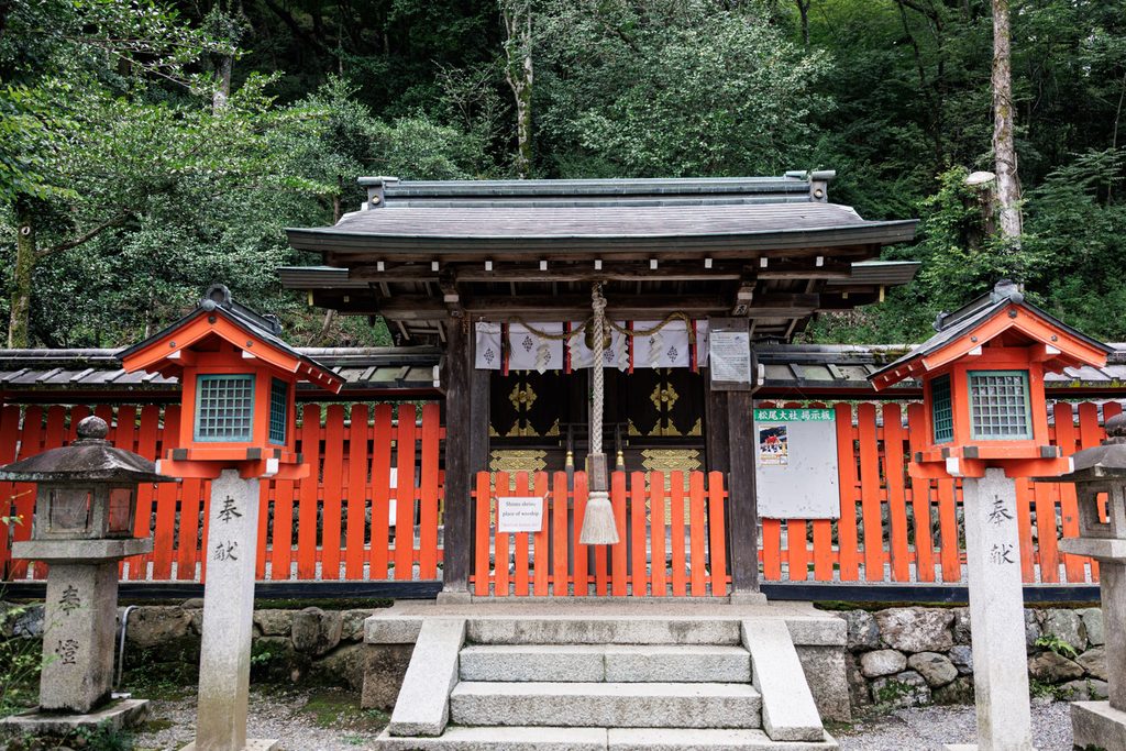 The main hall of Ichitani-Munakata Shrine in Arashiyama, Kyoto, behind a vermilion fence with stone lanterns in front