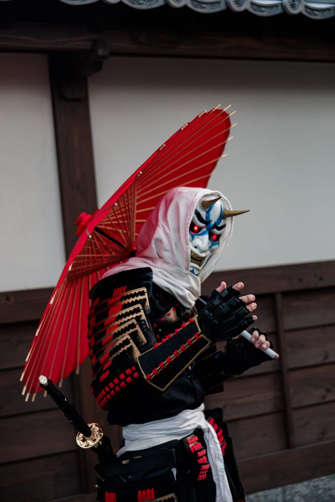Yokai performer in an oni mask and armor holding a red paper umbrella