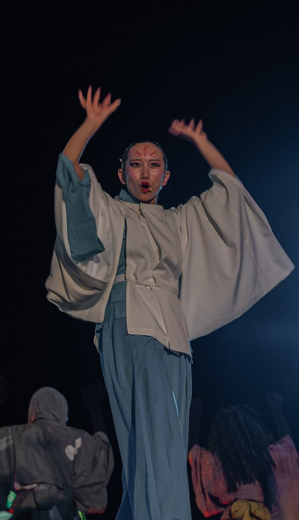 Stage dancer with red face paint raising both arms during the yokai show