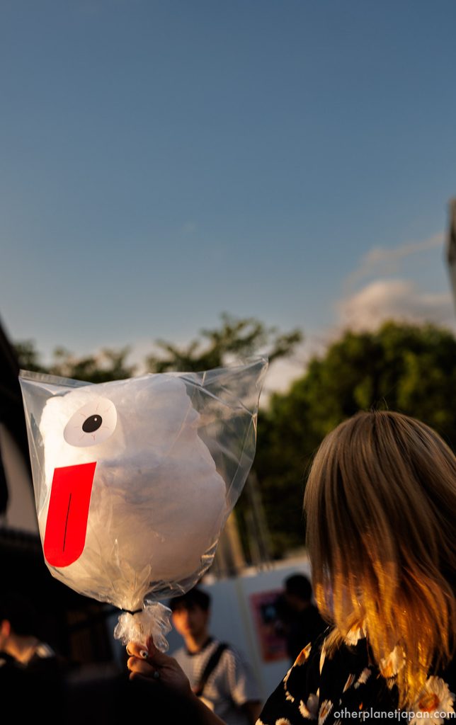 Yokai-themed cotton candy with one eye and a red tongue held up against the evening sky