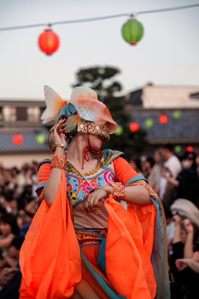 Dancer in a goldfish yokai headdress and orange robes under festival lanterns