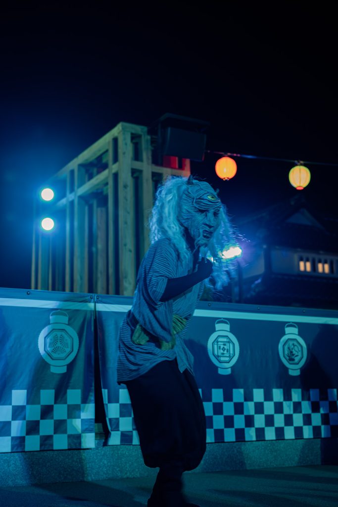One-eyed yokai with white hair dancing under blue light and paper lanterns at night