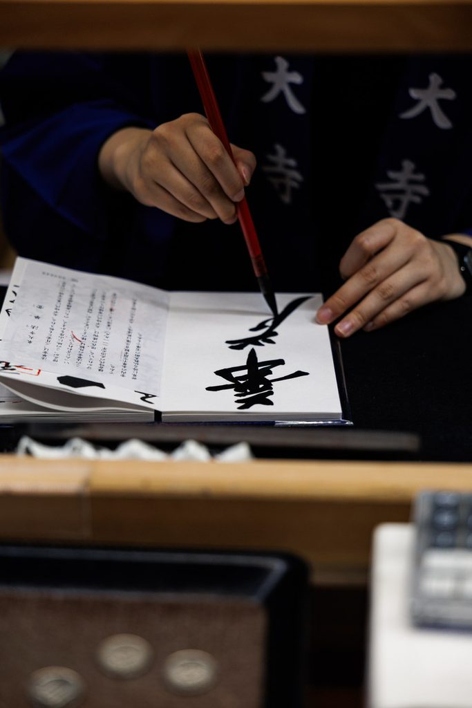 A Todaiji staff member in a temple happi coat writing the Kegon goshuin with a brush, coins on the counte