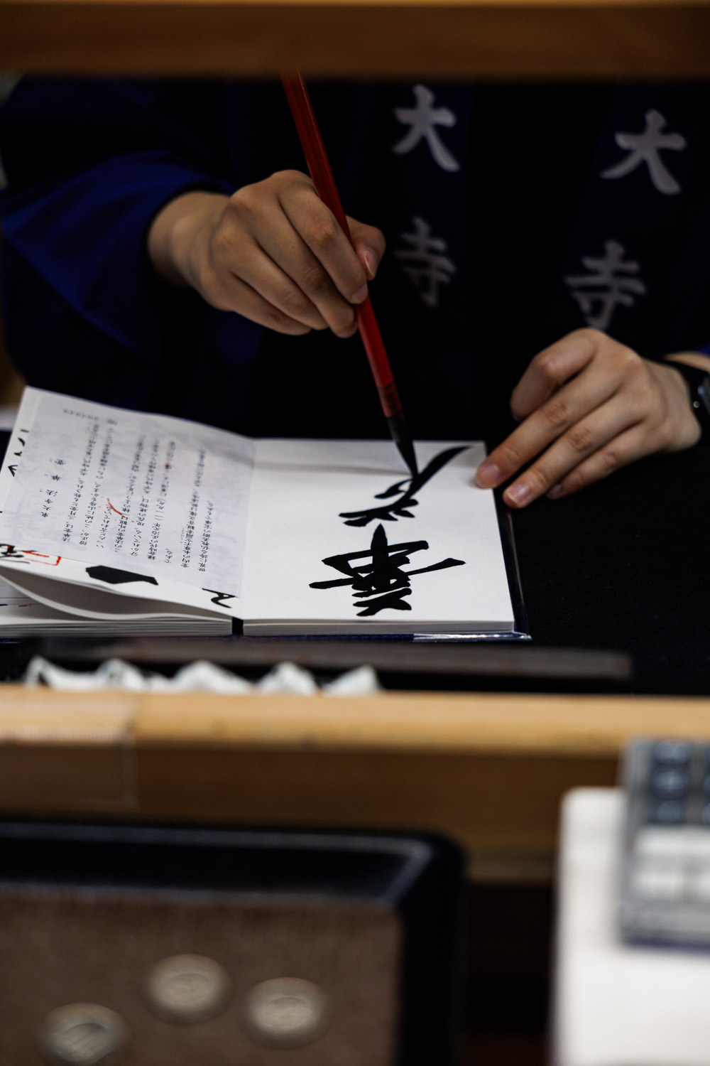 A Todaiji staff member in a temple happi coat writing the Kegon goshuin with a brush, coins on the counte
