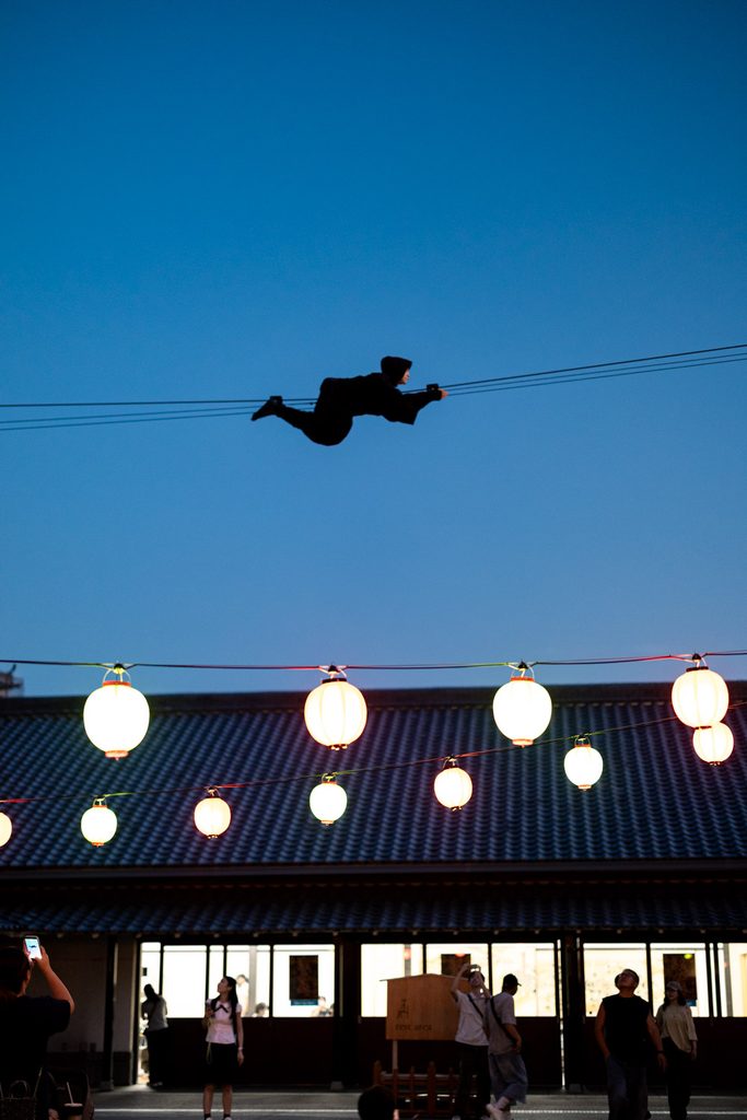 Ninja crossing a wire above glowing paper lanterns at dusk in Uzumasa, Kyoto
