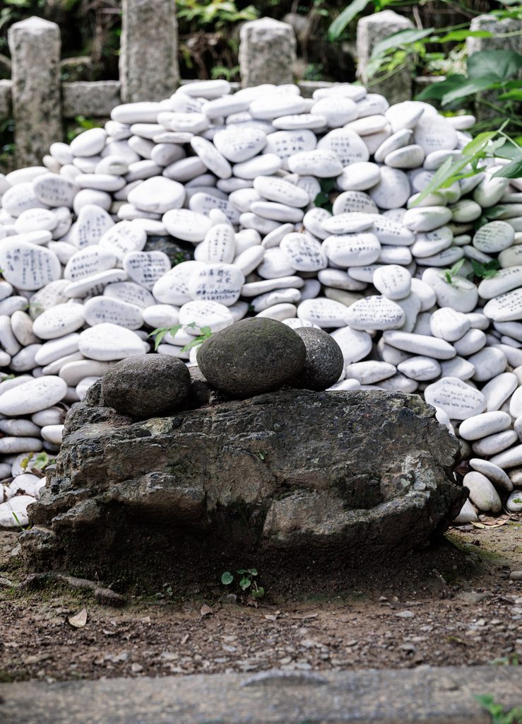 Tsukinobe-ishi, the dark birthing stone at Tsukiyomi Shrine in Kyoto, with hundreds of white prayer stones piled behind it